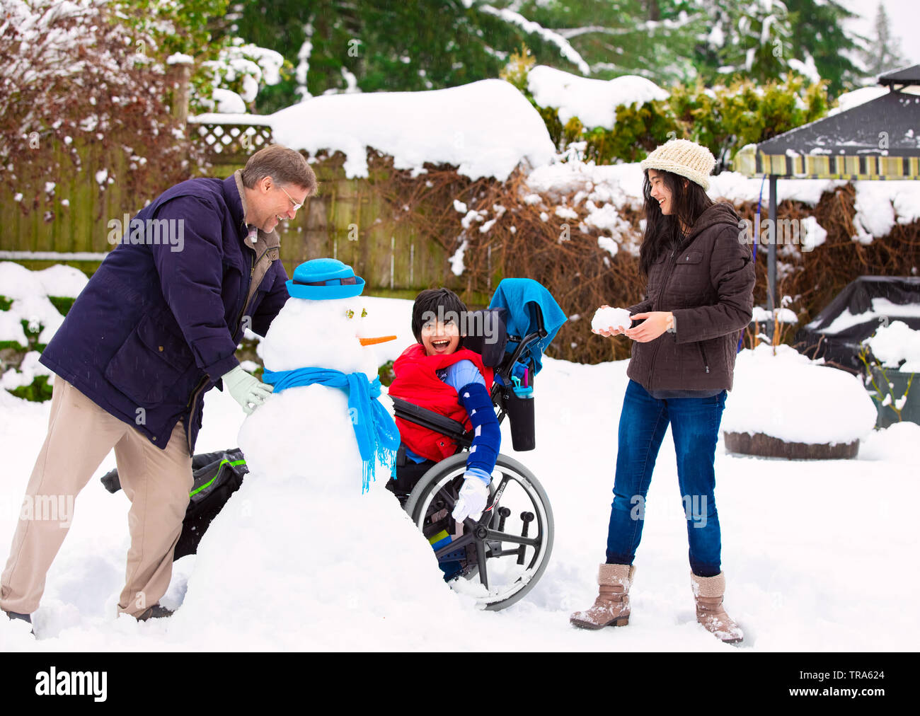 Disabled biracial young boy in wheelchair building a snowman with father and sister during winter after heavy snowfall Stock Photo