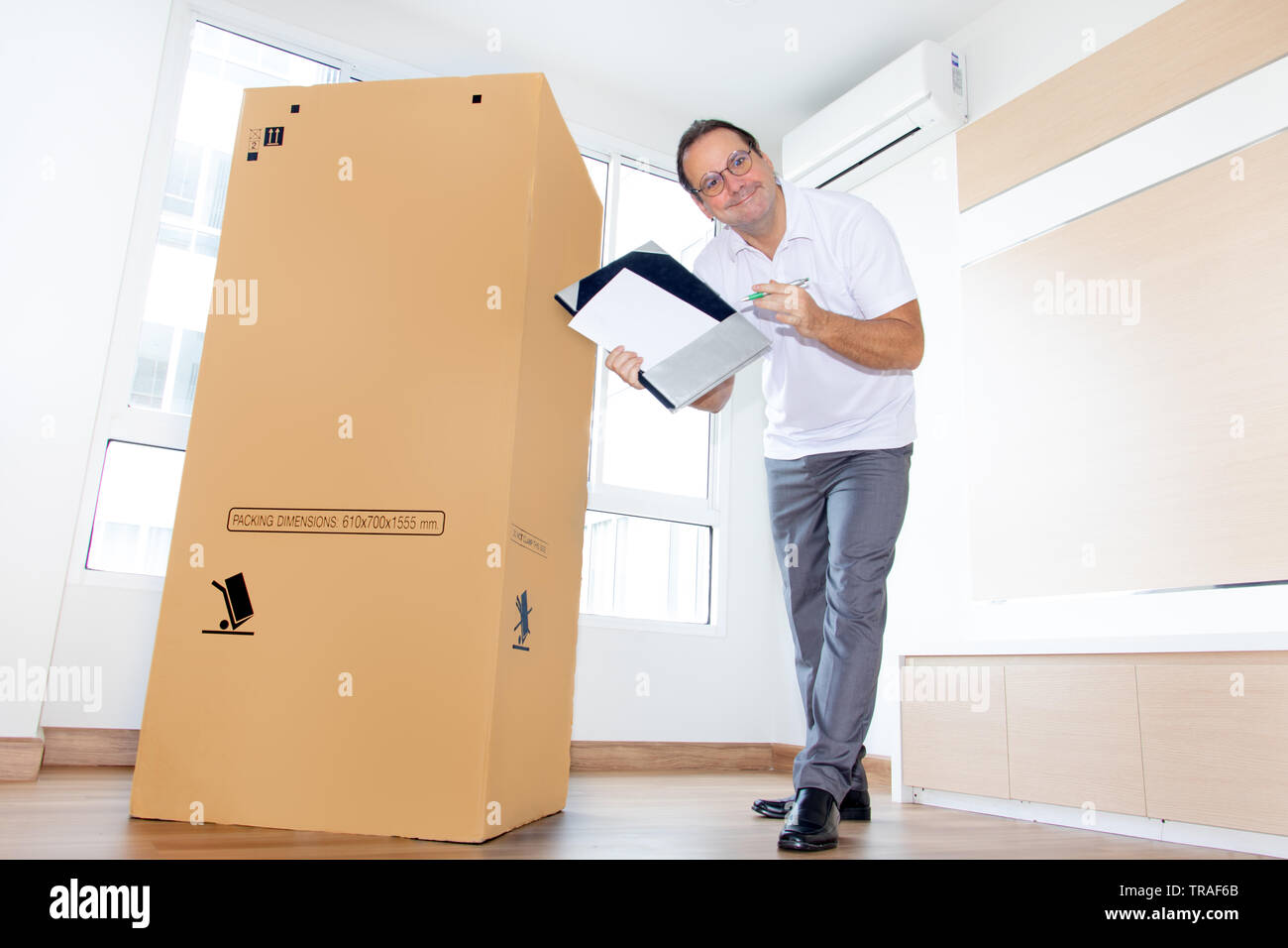 A man in an empty room shows a blank paper document for shipping large package. The postman delivers the parcel to the new apartment. Stock Photo