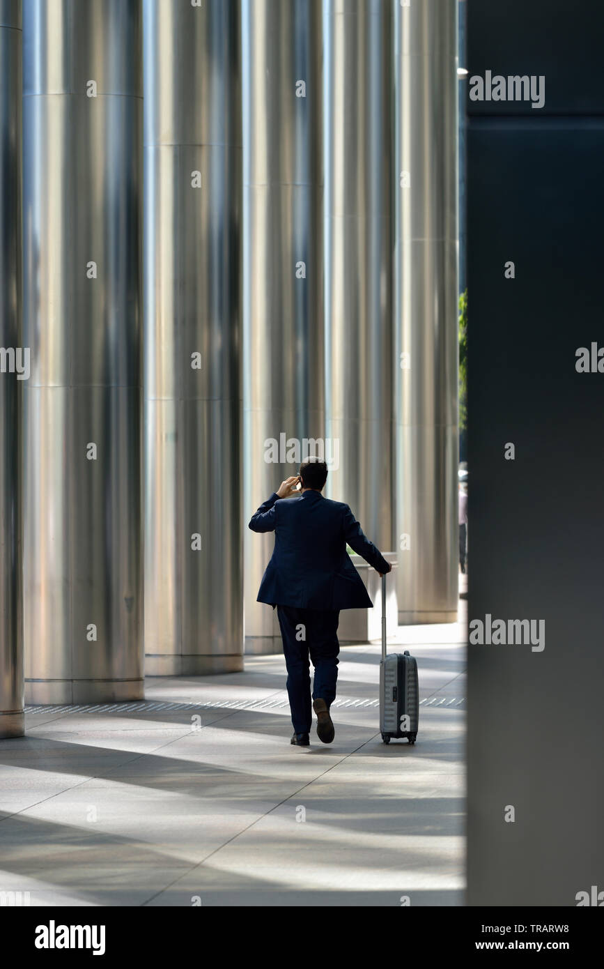 Business man in suit on mobile phone with roller case, Canary Wharf, London, United Kingdom Stock Photo