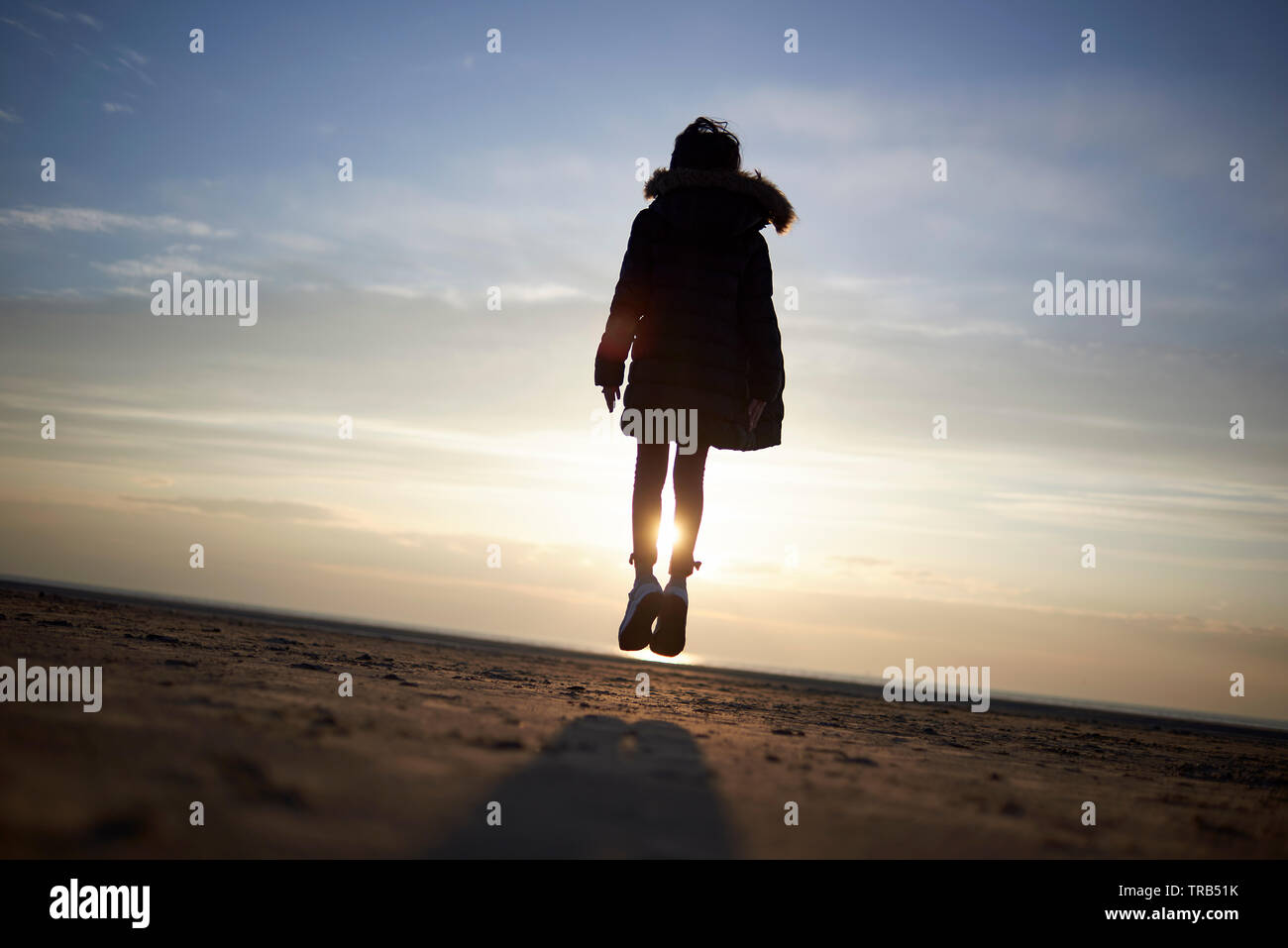Silhouette of a young girl jumping high in the air in a barren beach landscape making it look as if she is weightless and floating in the midair Stock Photo