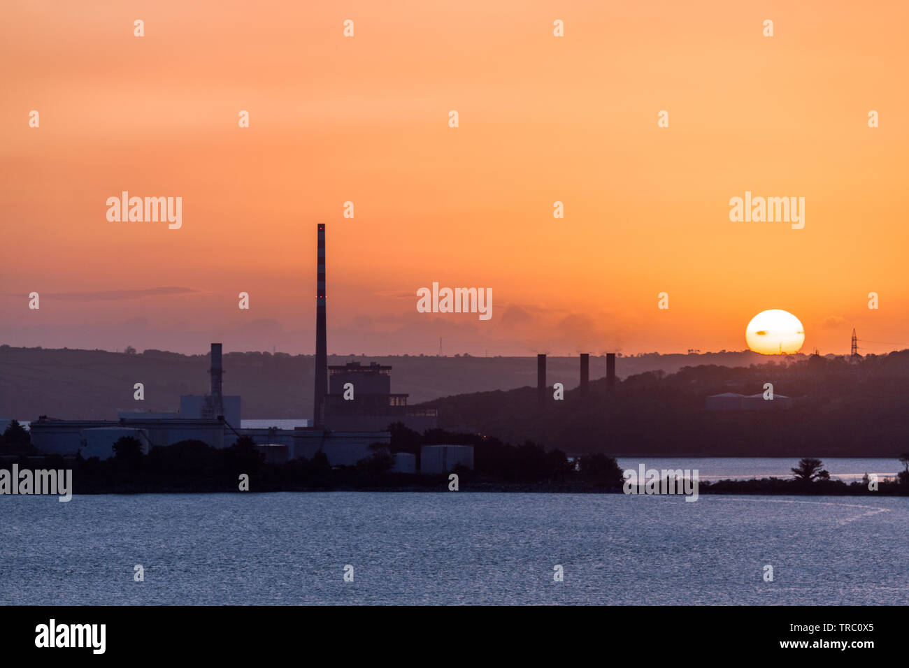 Aghada, Cork, Ireland. 03rd June, 2019. On a clear bank holiday Monday, the sun rises behind storage tanks at the oil refinery and the ESB generating station in Aghada, Co. Cork, Ireland. Credit; David Creedon / Alamy Live News Stock Photo