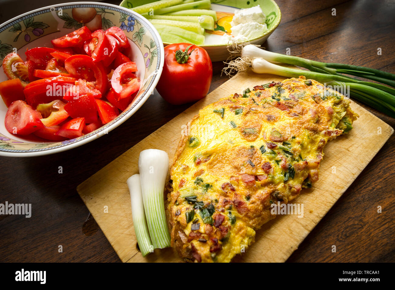 Delicious egg omlette with spring onion and bakon, served on a wooden plank and decorated with tomato salad, one tomato, cucumber, white cheese and sp Stock Photo