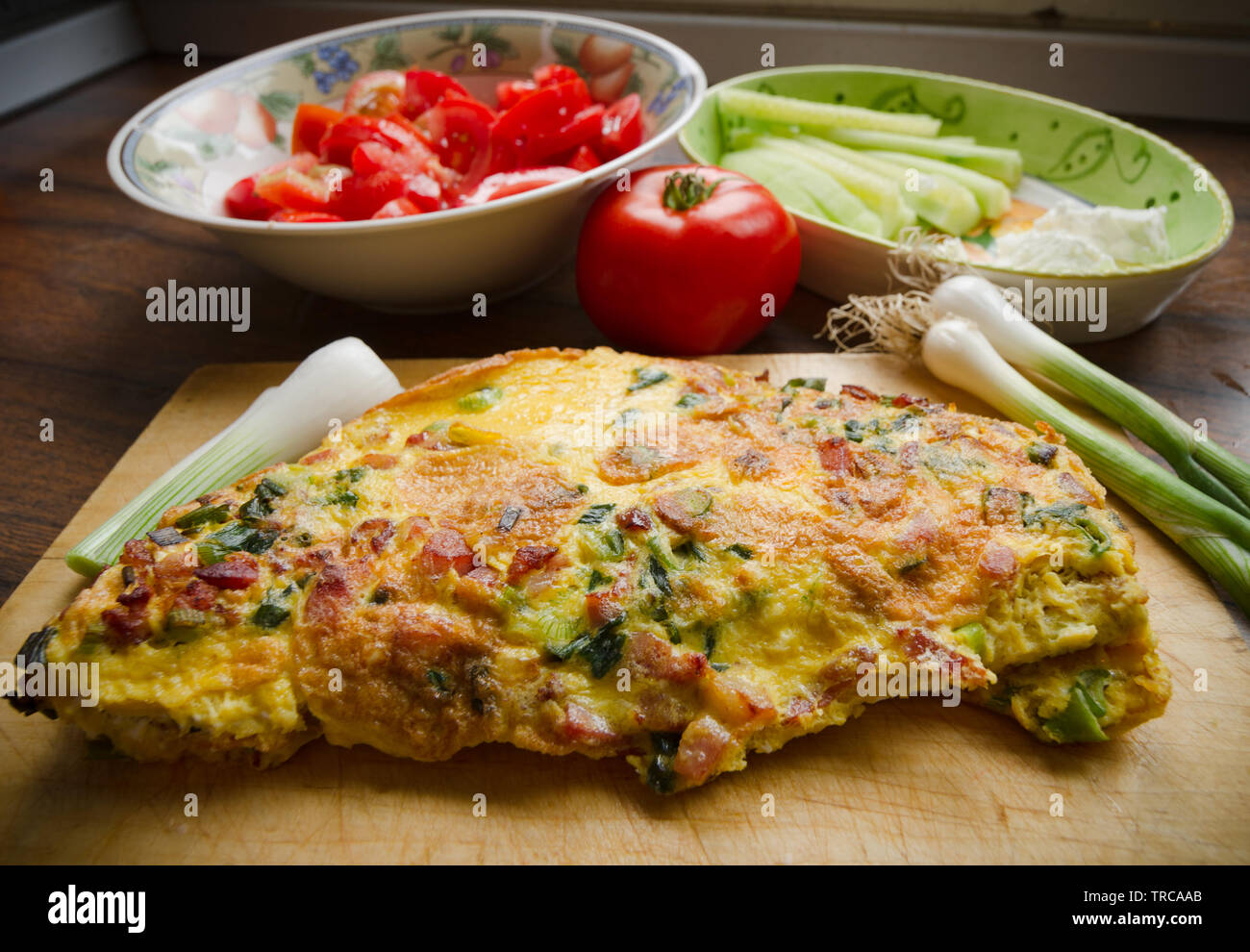 Delicious egg omlette with spring onion and bakon, served on a wooden plank and decorated with tomato salad, one tomato, cucumber, white cheese and sp Stock Photo