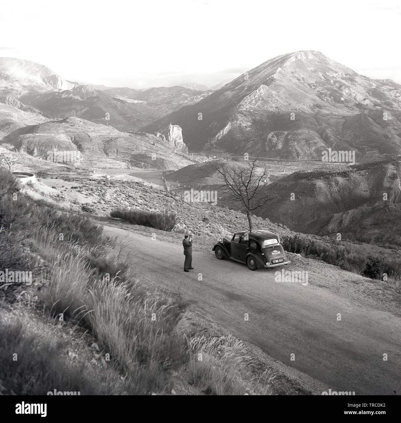 1950s, historical, a man outside filming the route of the Monte Carlo rally, with a car of the era parked on a road outside the French town of Sisteron, near the Alps.  A sign, 'Cinema Rallye Monte-Carlo' is on the back of the car, the numberplate - FWK 954 - a British car, with a GB sticker. Stock Photo