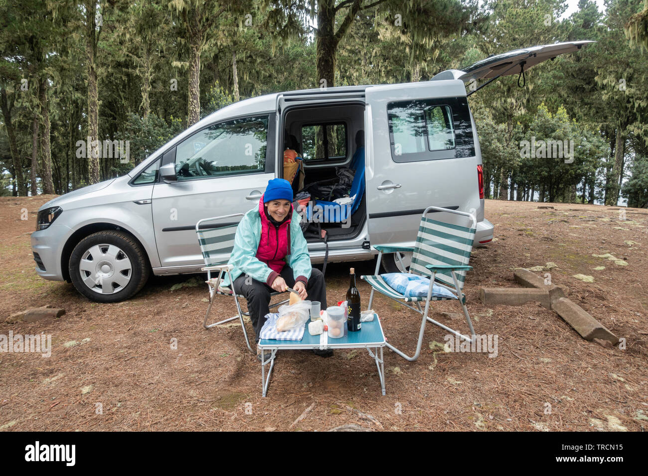 Woman with food and wine on picnic table next to small camper van in pine forest campsite in Spain. Stock Photo