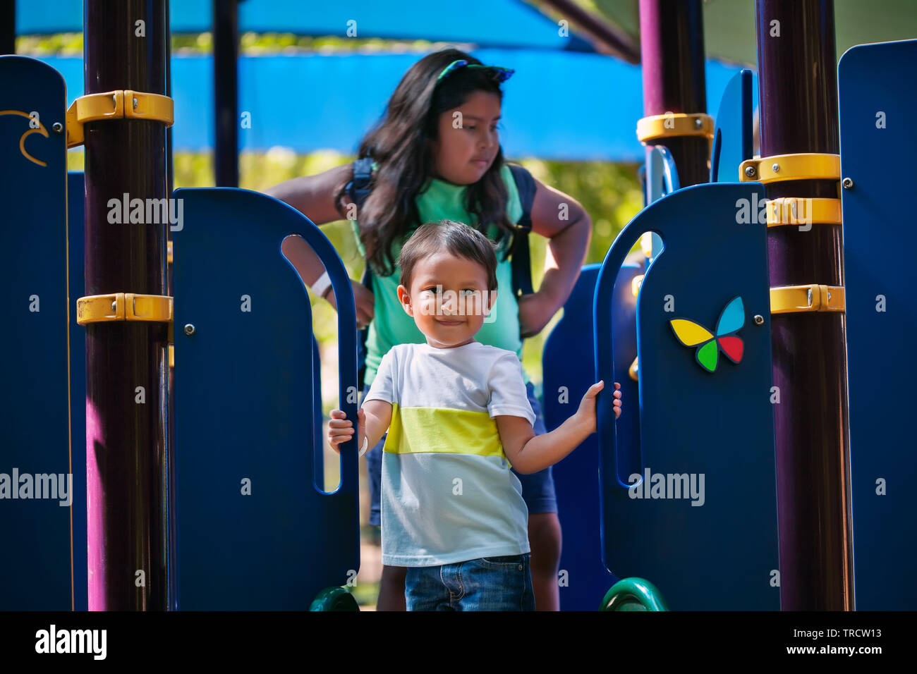 Young boy 3 years of age is standing and holding on to playground slide with older sister standing behind to help. Stock Photo