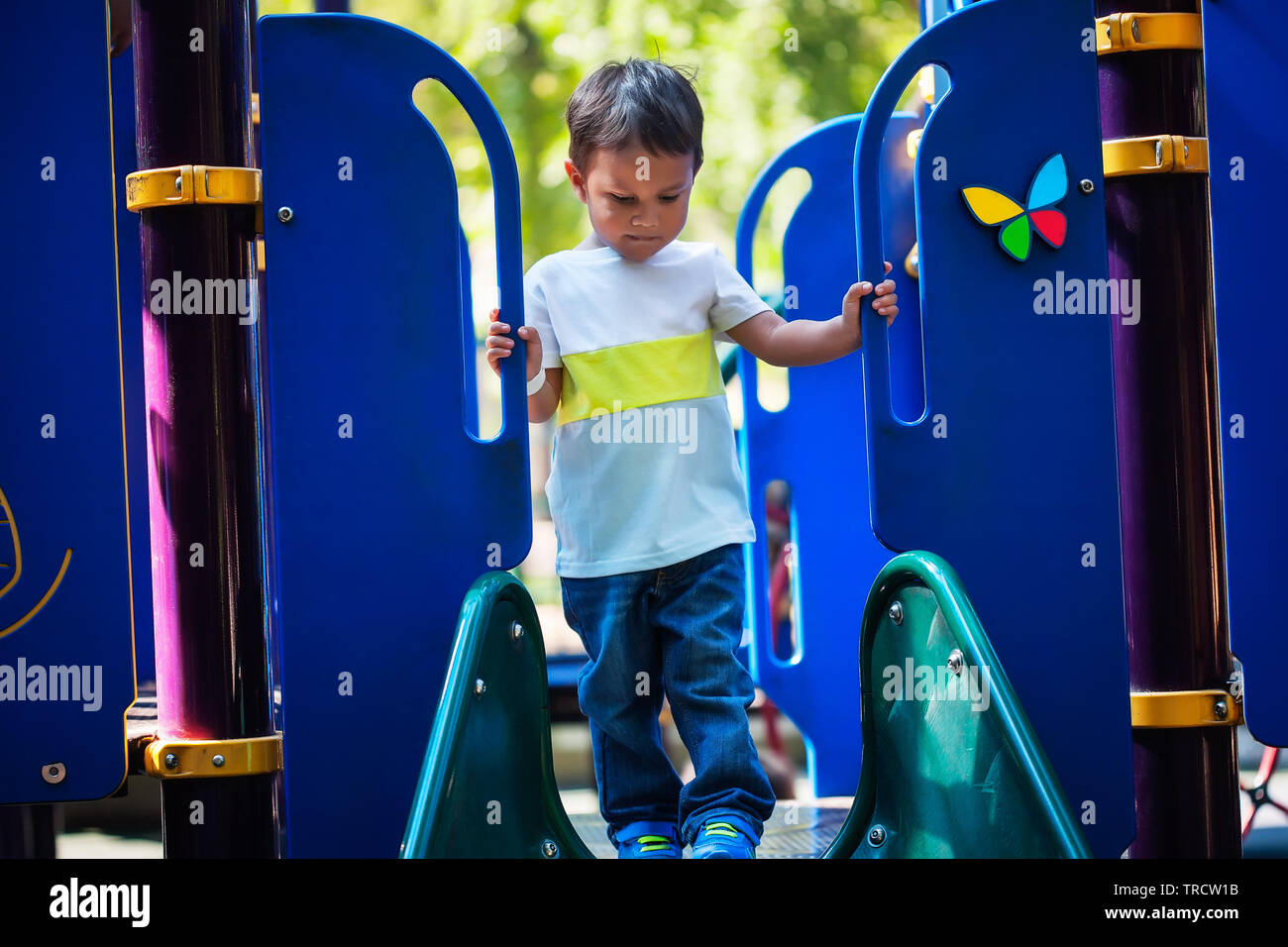 3 year old boy looks unsure about going down a slide as he looks down at the edge of the playground slide. Stock Photo