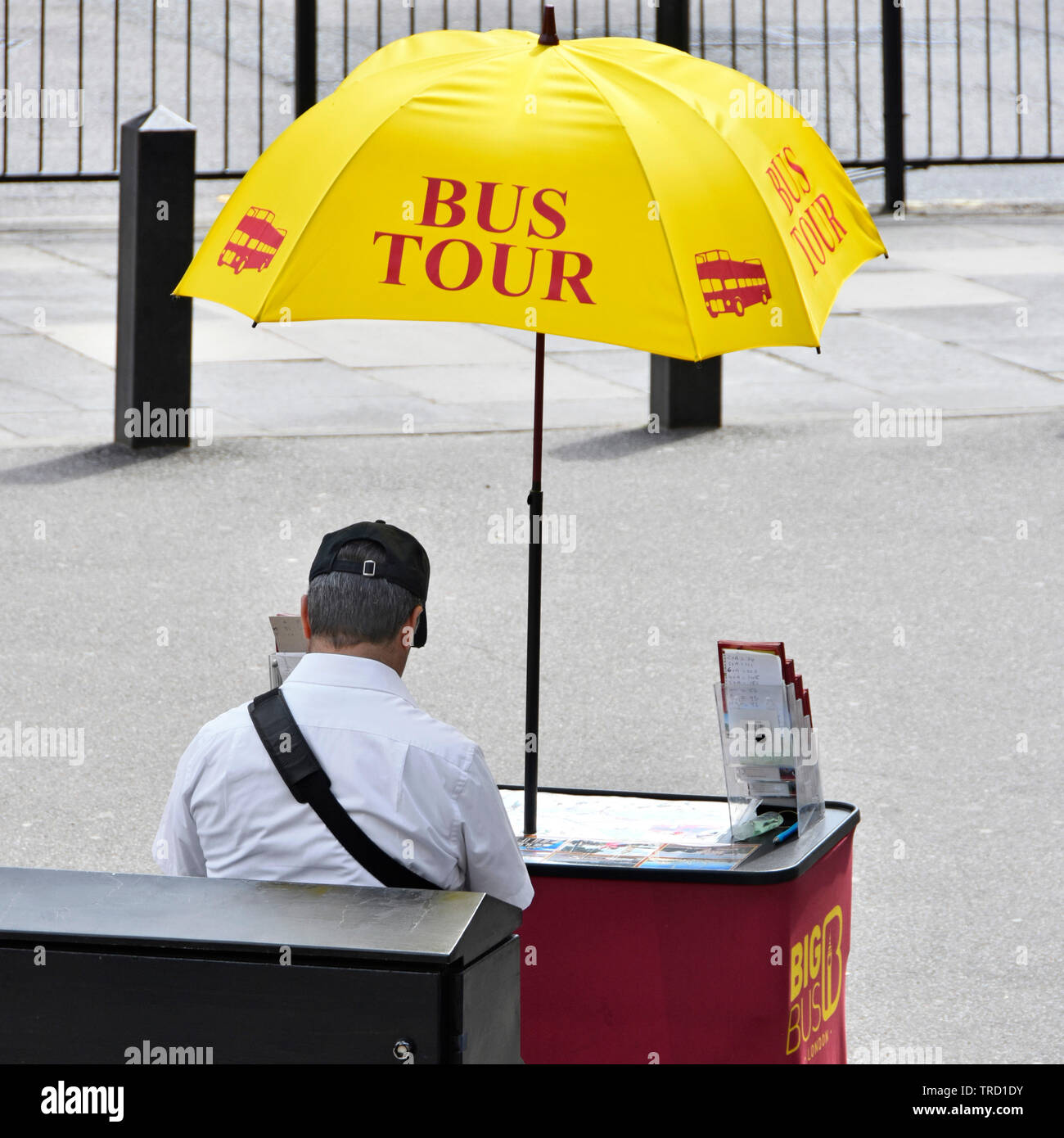 London street scene tourism tour bus salesman sitting on pavement at small desk under yellow advertising umbrella hot summer day Westminster Abbey UK Stock Photo