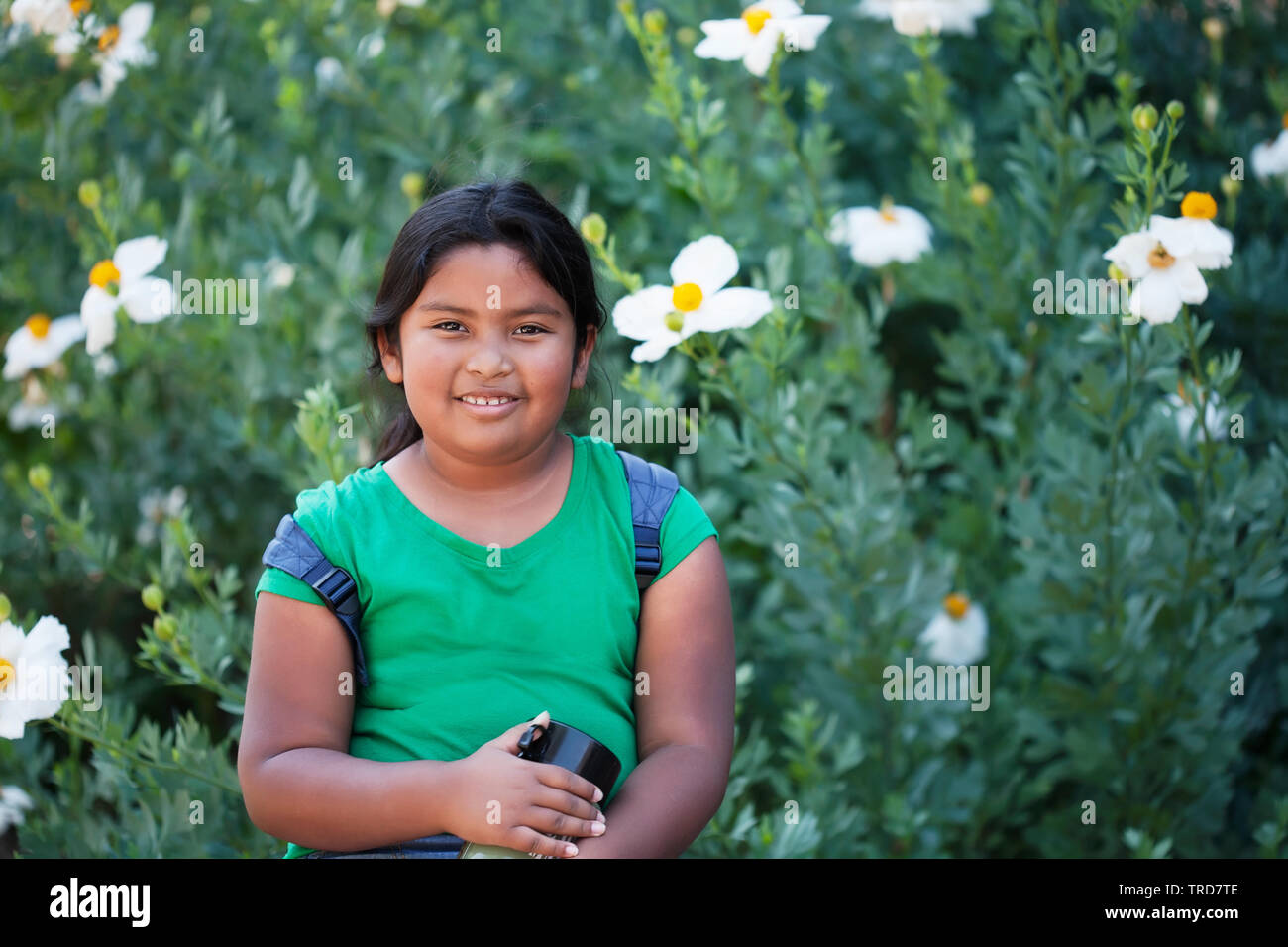 A 9 year old child or student wearing a backpack and holding a thermos standing in front of blooming flowers during a school field trip. Stock Photo
