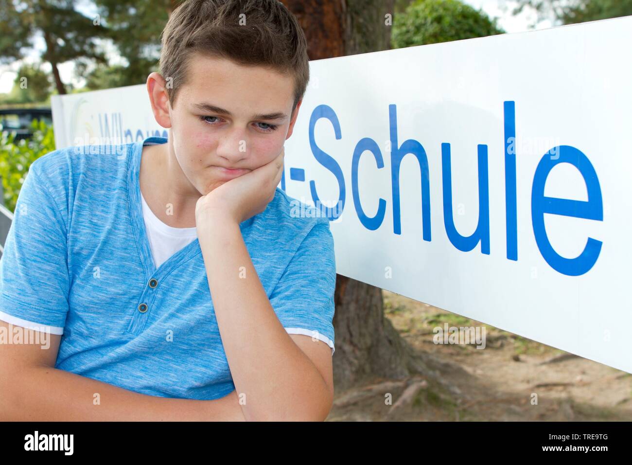 Angry looking boy sitting outside in front of a school sign Stock Photo