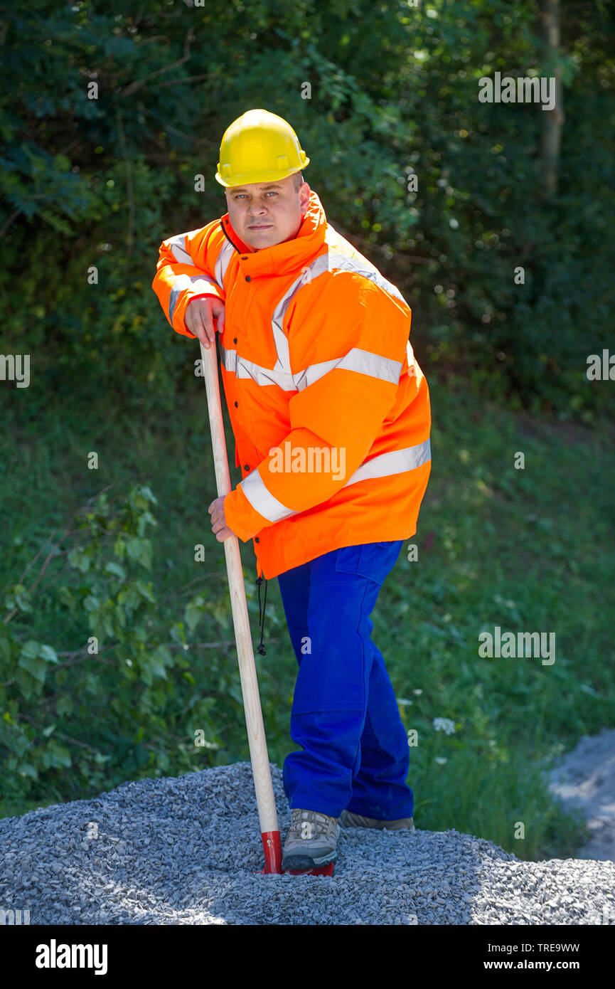 Construction worker in a orange high-visibility jacket, shoveling pebble stones next to a road Stock Photo