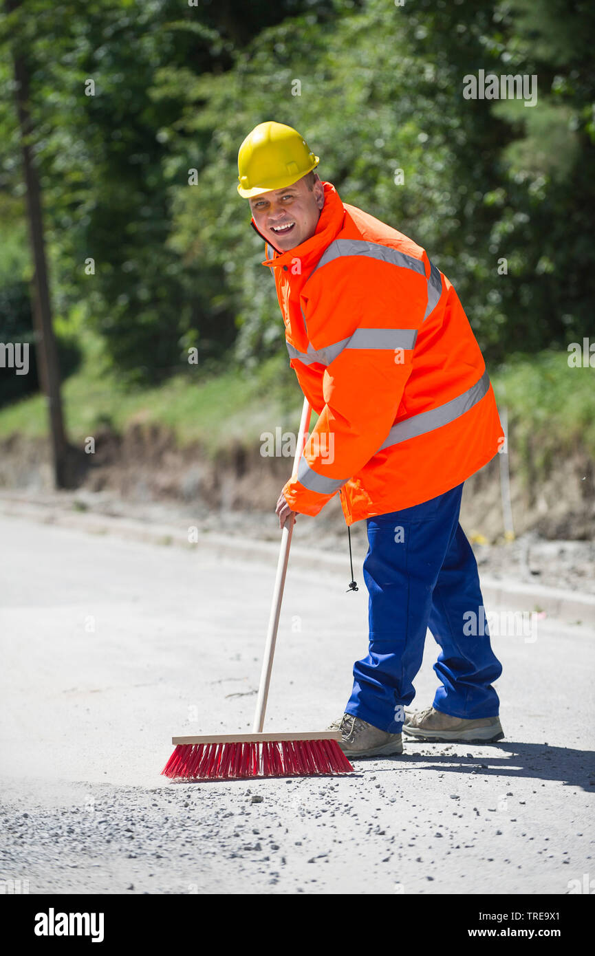 Construction worker in a orange high-visibility jacket, sweeping pebble stones on a road Stock Photo