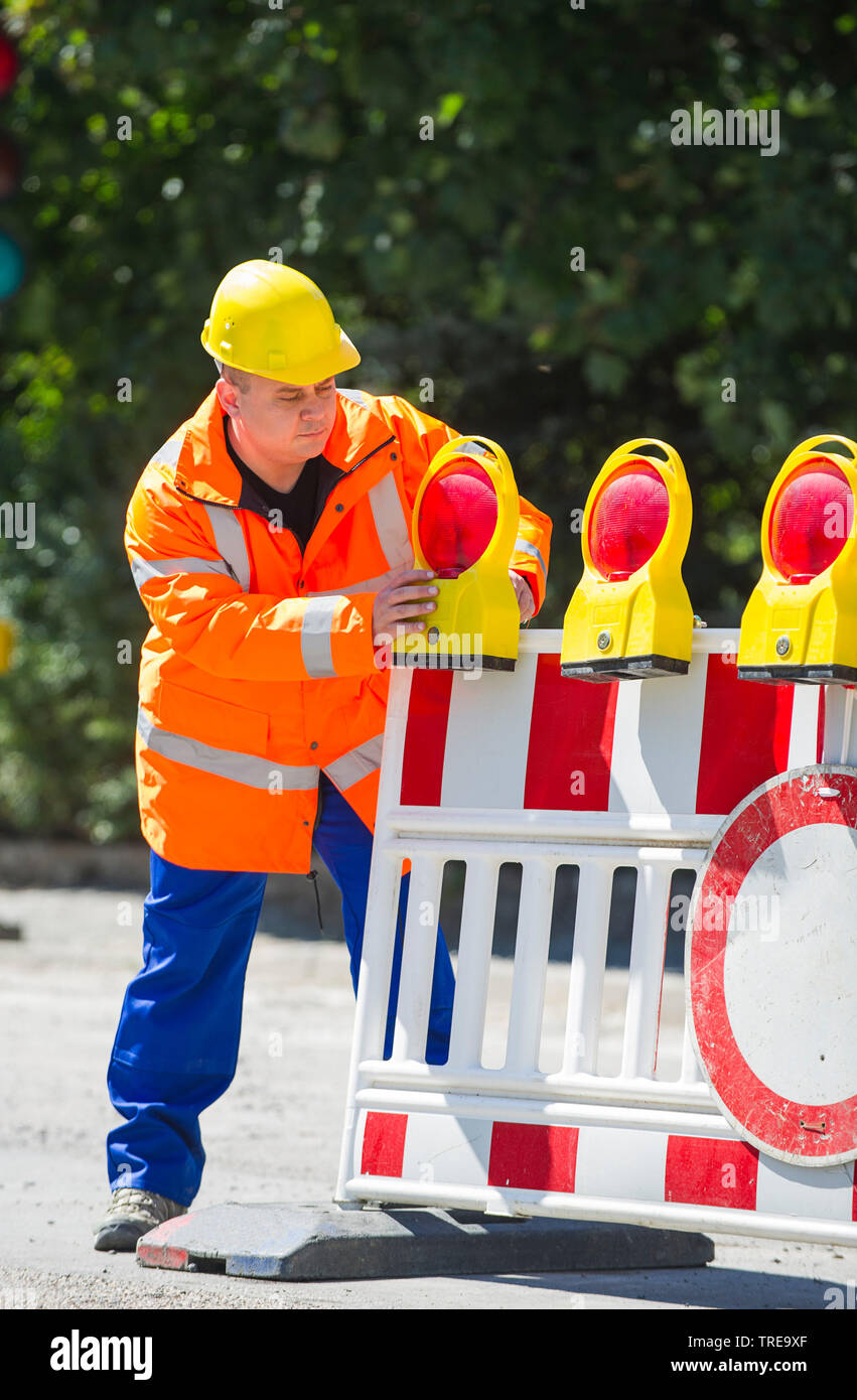 Construction worker in a orange high-visibility jacket, installing a roadblock with construction warning lights Stock Photo