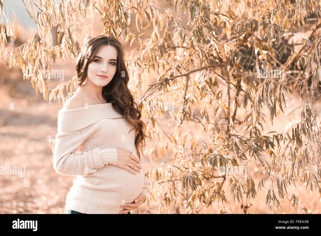 Stylish pregnant girl 20-24 year old wearing cozy knitted sweater holding tummy outdoors. Looking at camera. Motherhood. Maternity. 20s. Stock Photo
