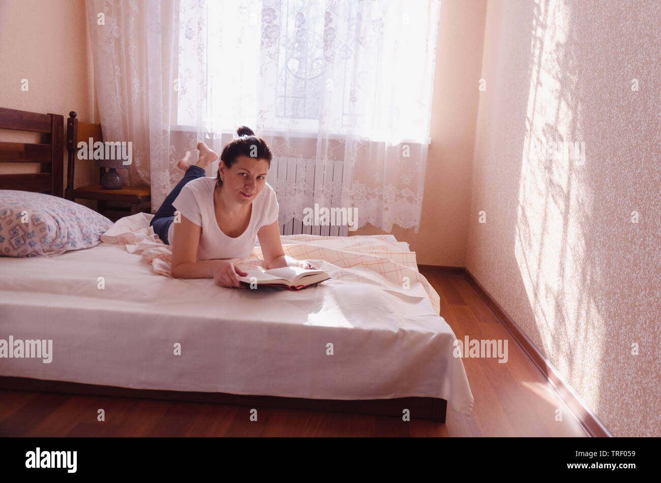 A happy young woman reading a book in a cozy bedroom in the sunny morning. Stock Photo