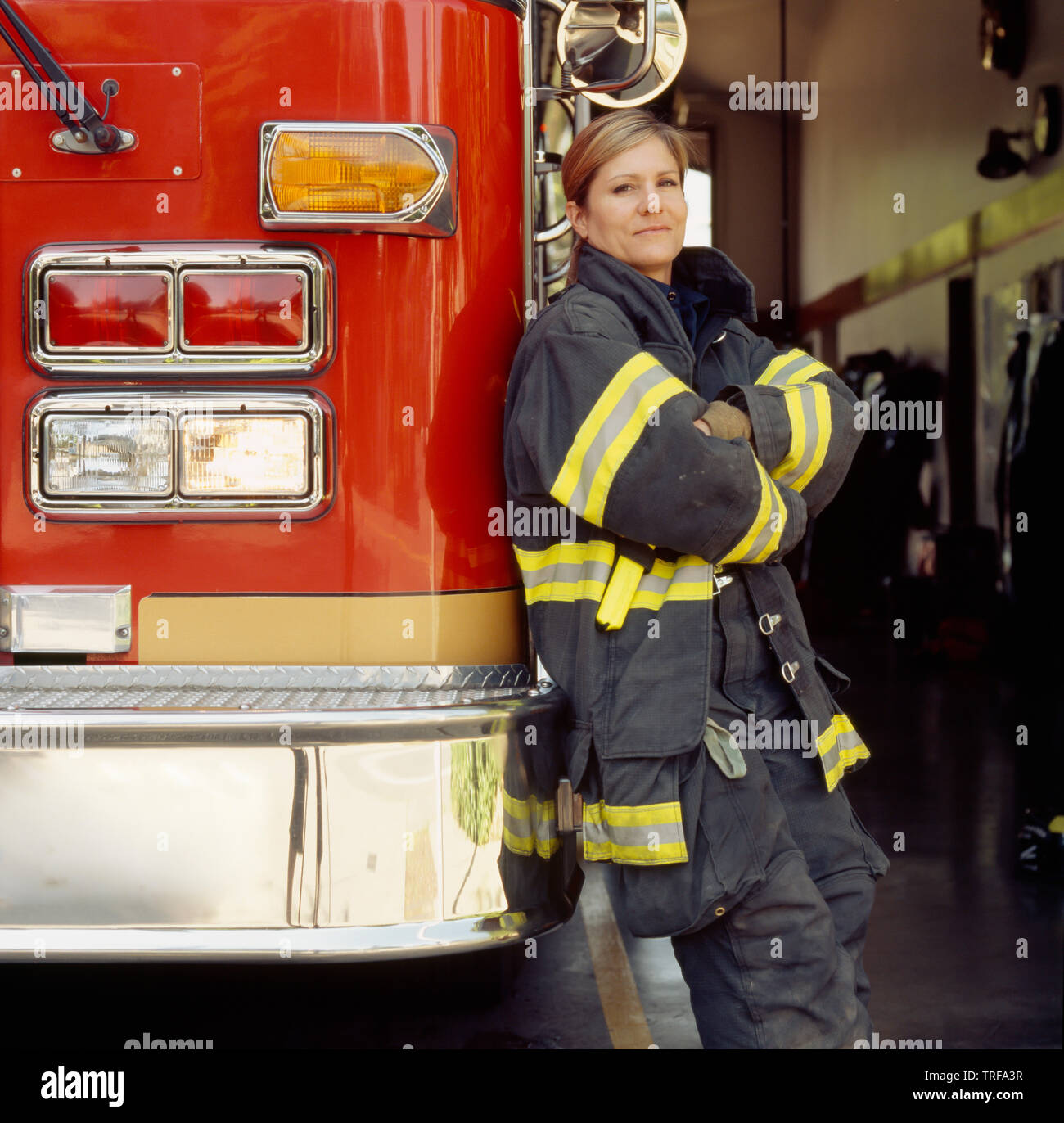 Smiling female woman firefighter wearing technical gear with fire engine truck at station. Confident, successful public service career professional. Stock Photo