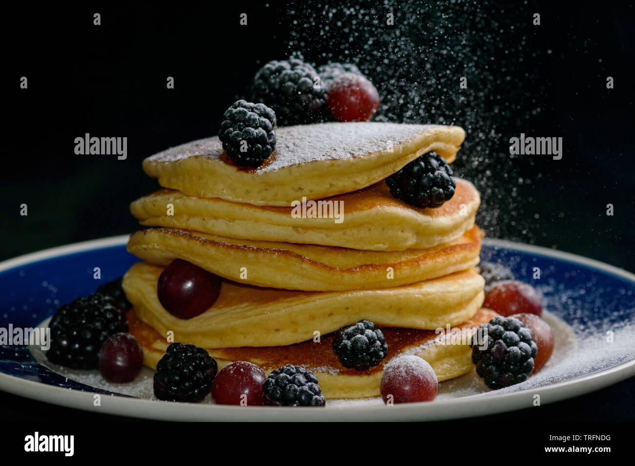 Stacked pancakes with summer berries and falling confectioners sugar , isolated on black. Stock Photo
