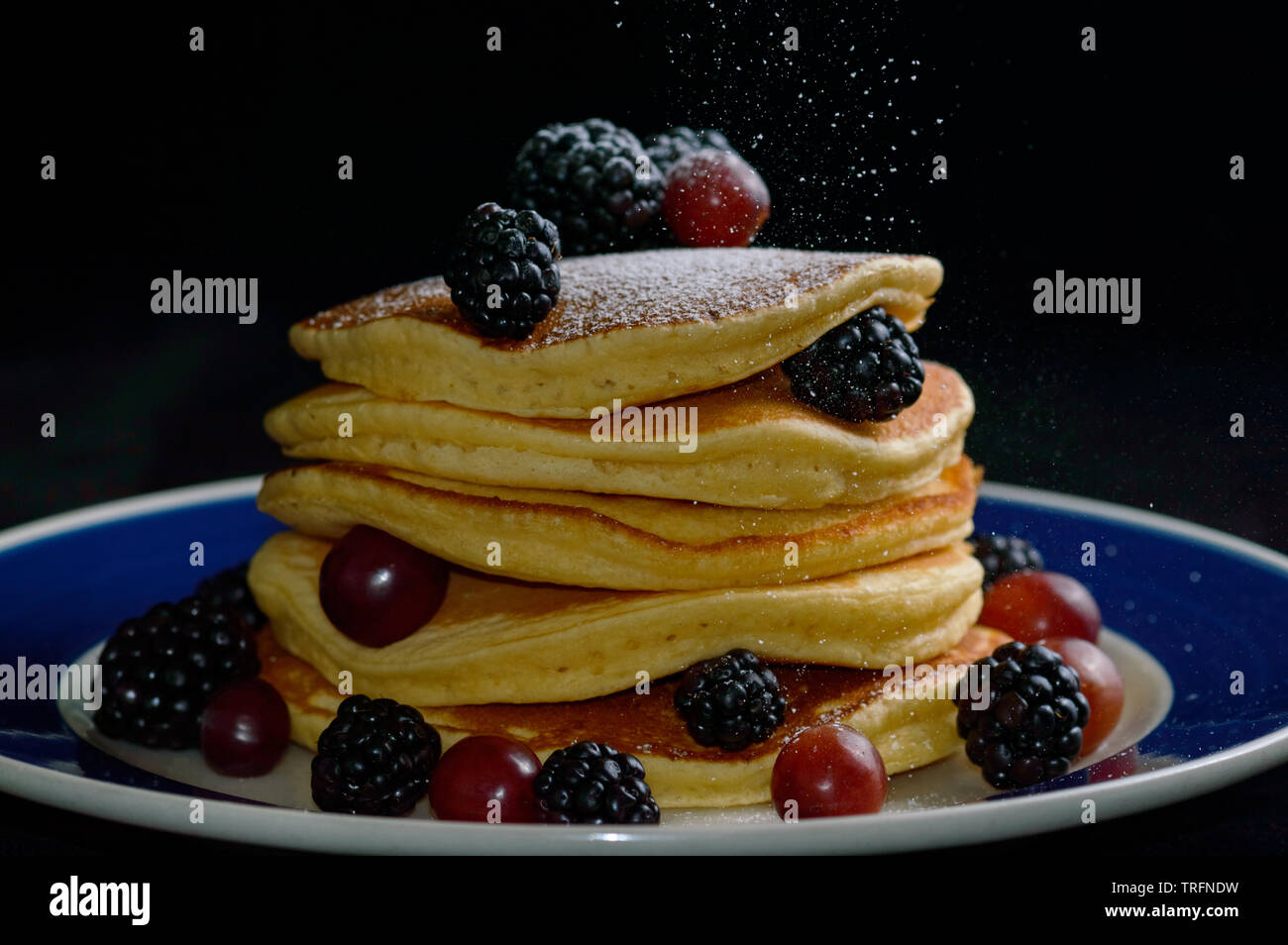 Stacked pancakes with summer berries and falling powdered sugar, isolated on black. Stock Photo