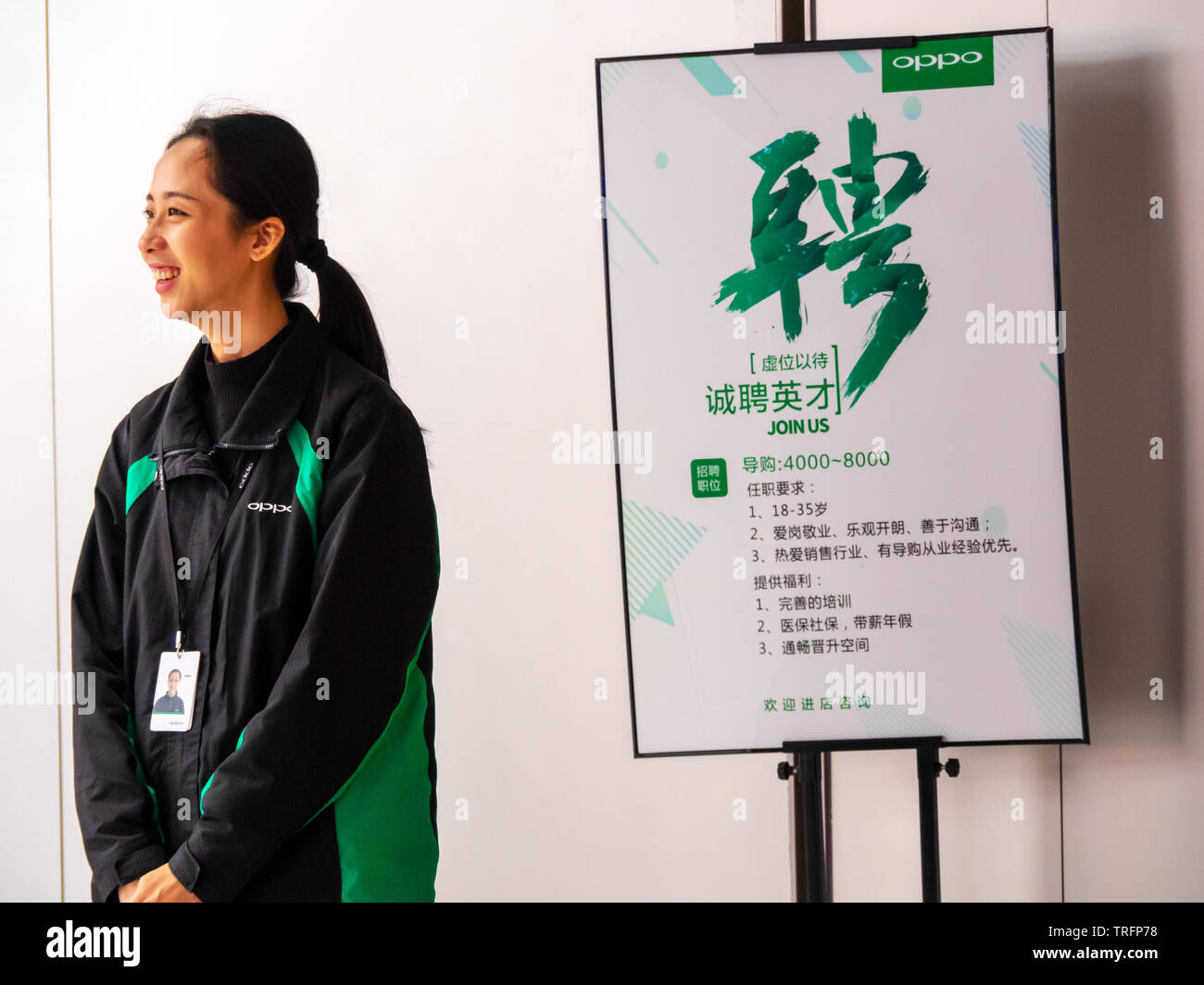 TIANHE, GUANGZHOU CITY, CHINA - 7 MAR 2019 - Smiling young Asian Chinese female Oppo employee stands next to an staff recruitment poster Stock Photo