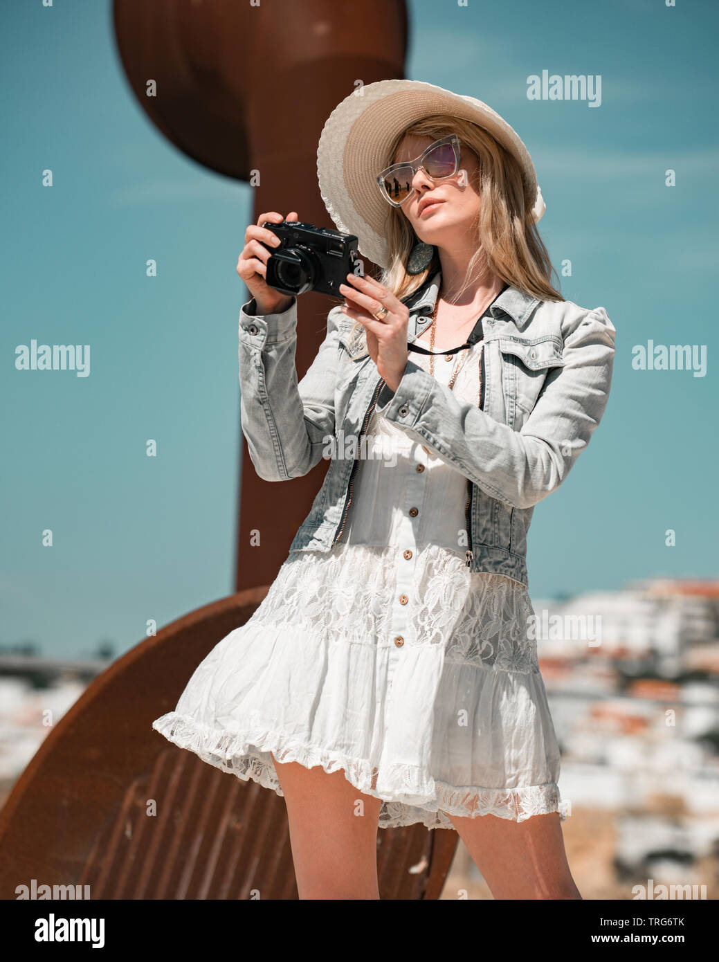 Blonde woman wearing a white dress and denim jacket in Portugal holding a Fuji camera wearing sunglasses. Stock Photo