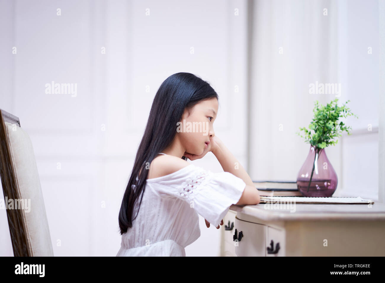 sad little asian girl with long black hair sitting at desk in her room thinking with hand on chin Stock Photo