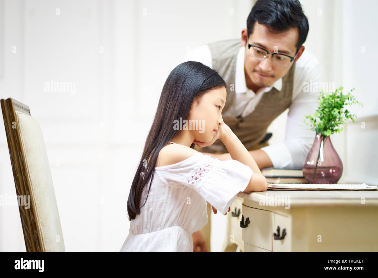 sad little asian girl sitting at desk in her room and getting comfort from caring father Stock Photo