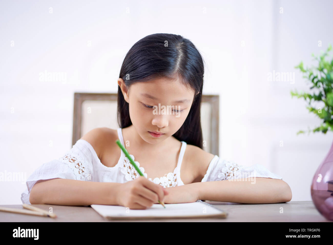 beautiful little asian girl with long black hair sitting at desk in her room writing or drawing on note book Stock Photo