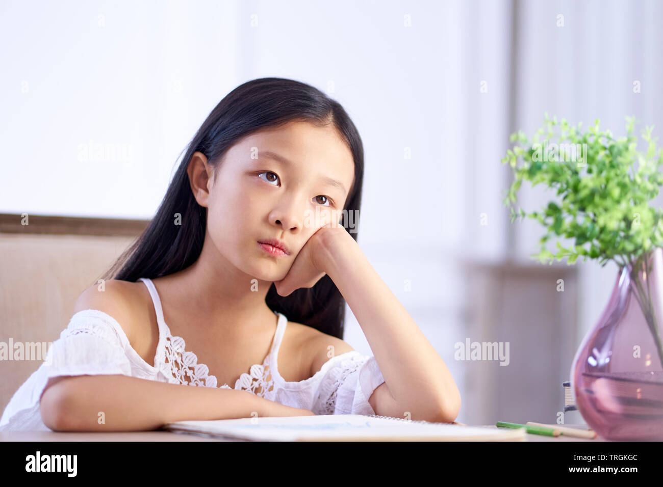 beautiful little asian girl with long black hair sitting at desk in her room looking up thinking. Stock Photo