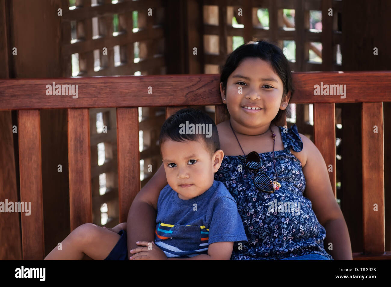 Little brother sitting next to his older sister in a wooden bench. Stock Photo
