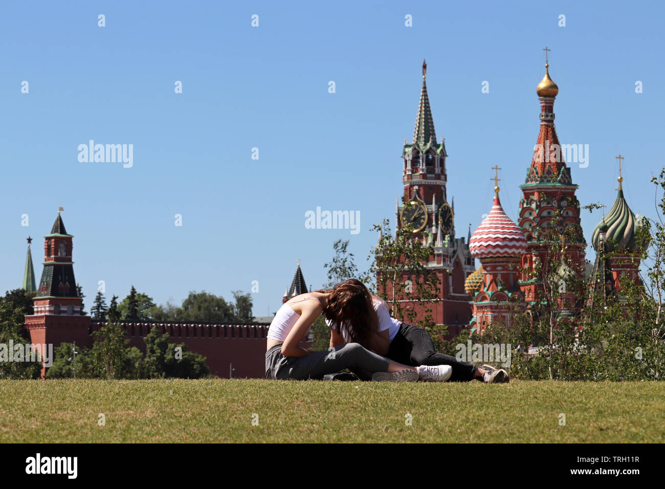 Girls sitting on a green grass in the Zaryadye park on background of Kremlin towers and St. Basil's Cathedral. Hot weather in Moscow, summer leisure Stock Photo