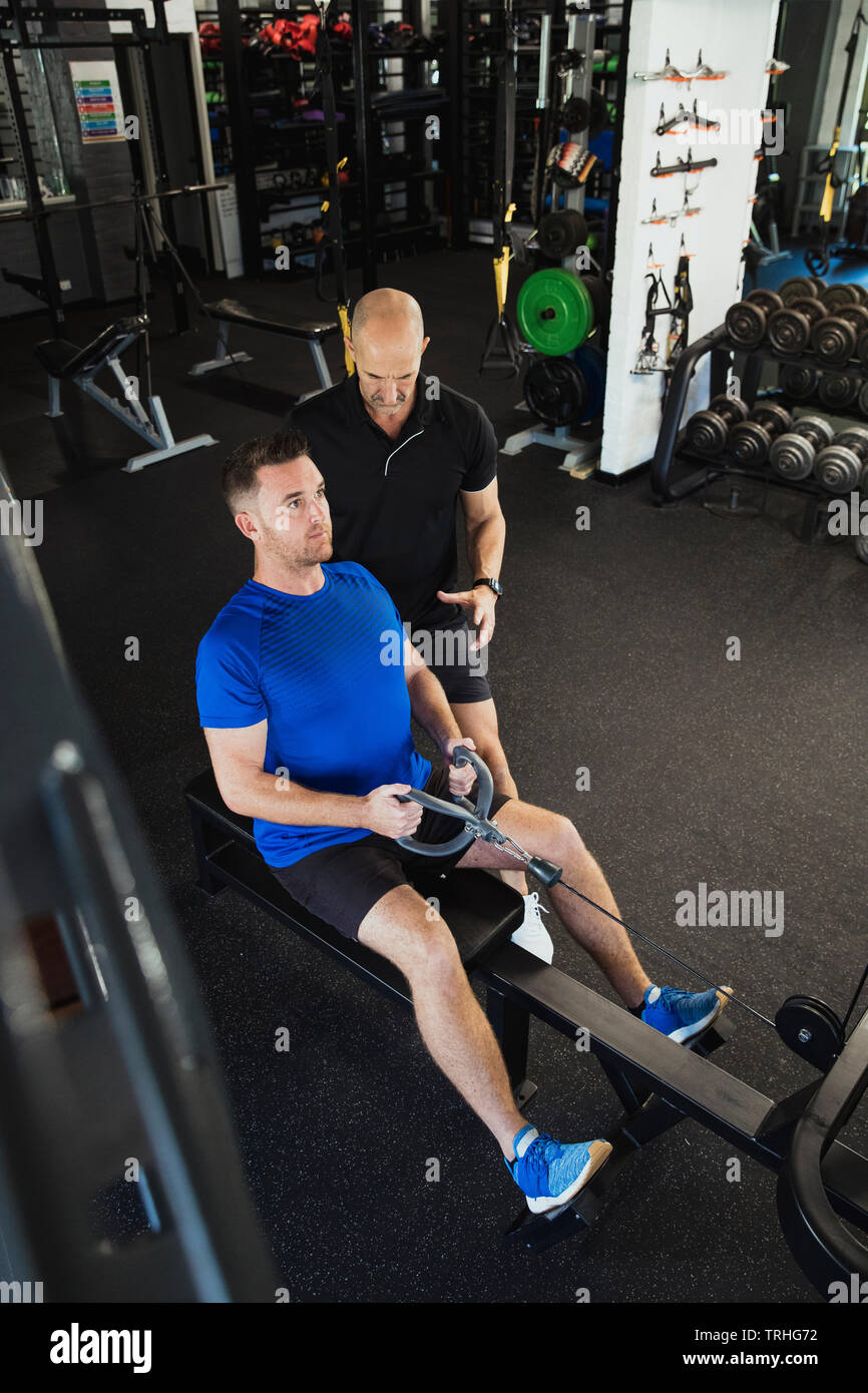 A  high-angle view of a mid-adult caucasian man training in the gym, he is using a rowing machine. A fitness instructor can be seen standing by the ma Stock Photo