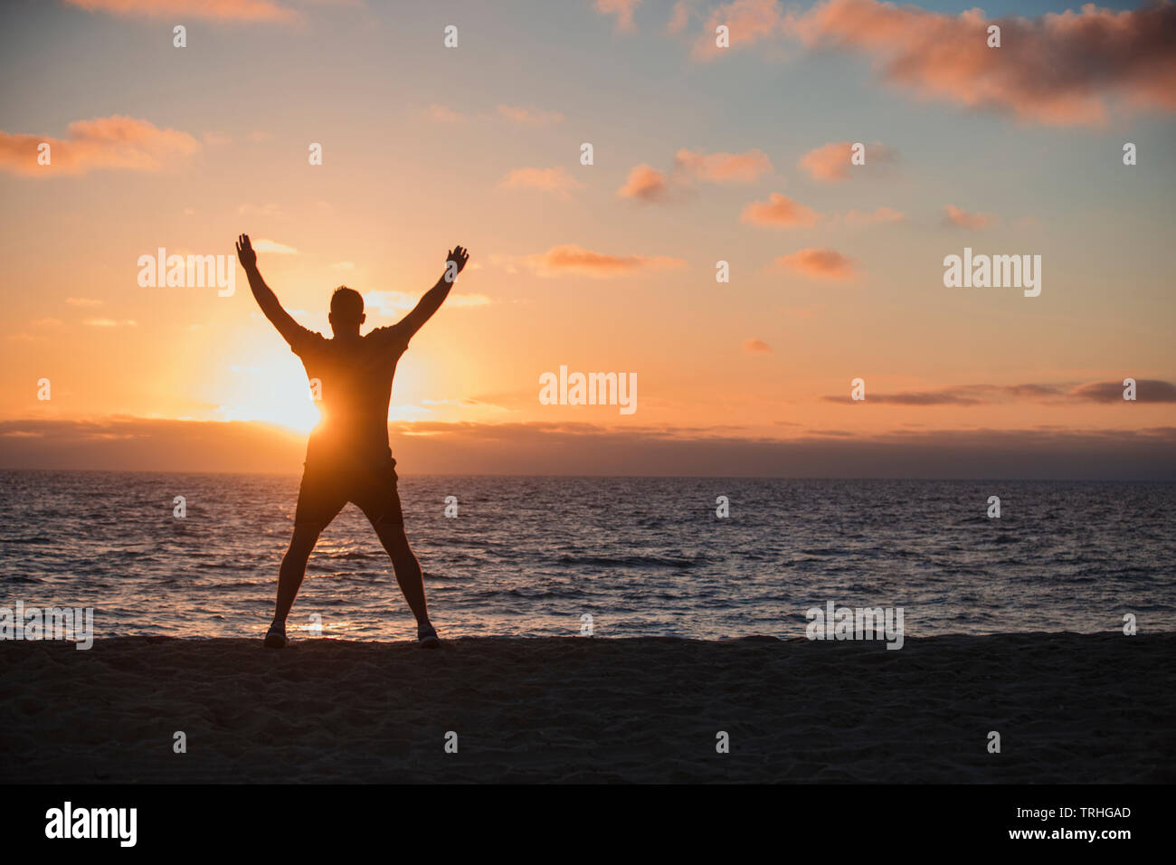 A rear-view shot of an unrecognizable man doing jumping jacks on the beach, he is looking out towards the sea and the sunset on a beautiful evening in Stock Photo