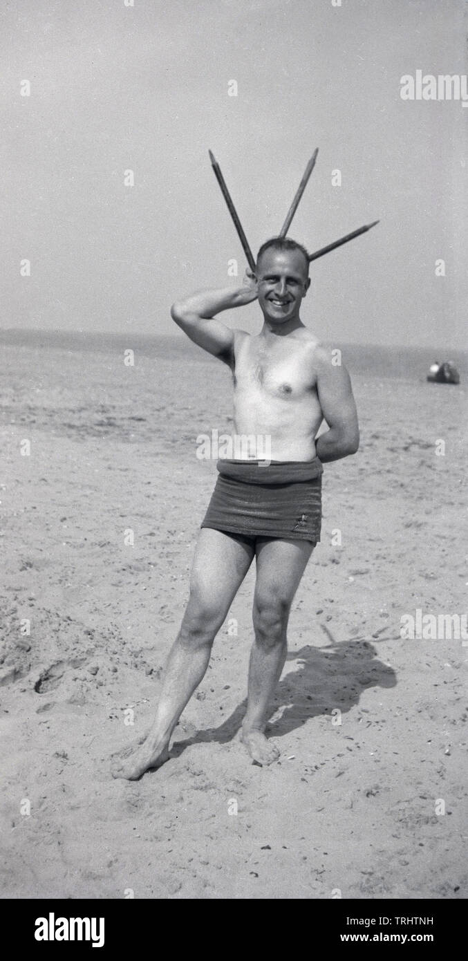 1930s, historical, man on a beach in a rolle down one-piece swimsuit of the era holding three cricket stumps over his head in the shape of a star, England, UK. Stock Photo