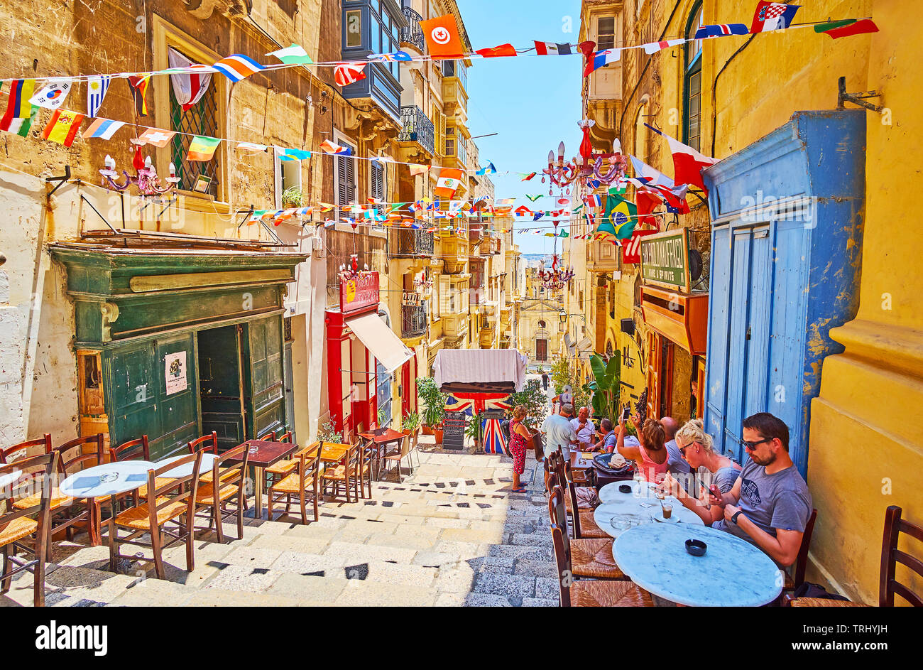 VALLETTA, MALTA - JUNE 19, 2018: The tables of cozy outdoor bar stretch along the descent of St Lucia street, decorated with colorful flag garlands, o Stock Photo
