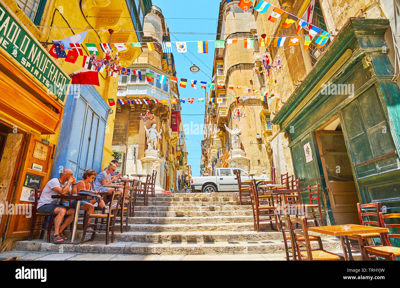 VALLETTA, MALTA - JUNE 19, 2018: The narrow descent of St Lucia street is lined with tables of local bars and cafes, decorated with flag garlands, on Stock Photo