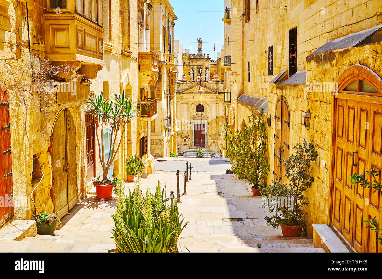 The medieval St Lucia street is decorated with green plants in pots; the same named church is seen at the end of the descent, Valletta, Malta. Stock Photo