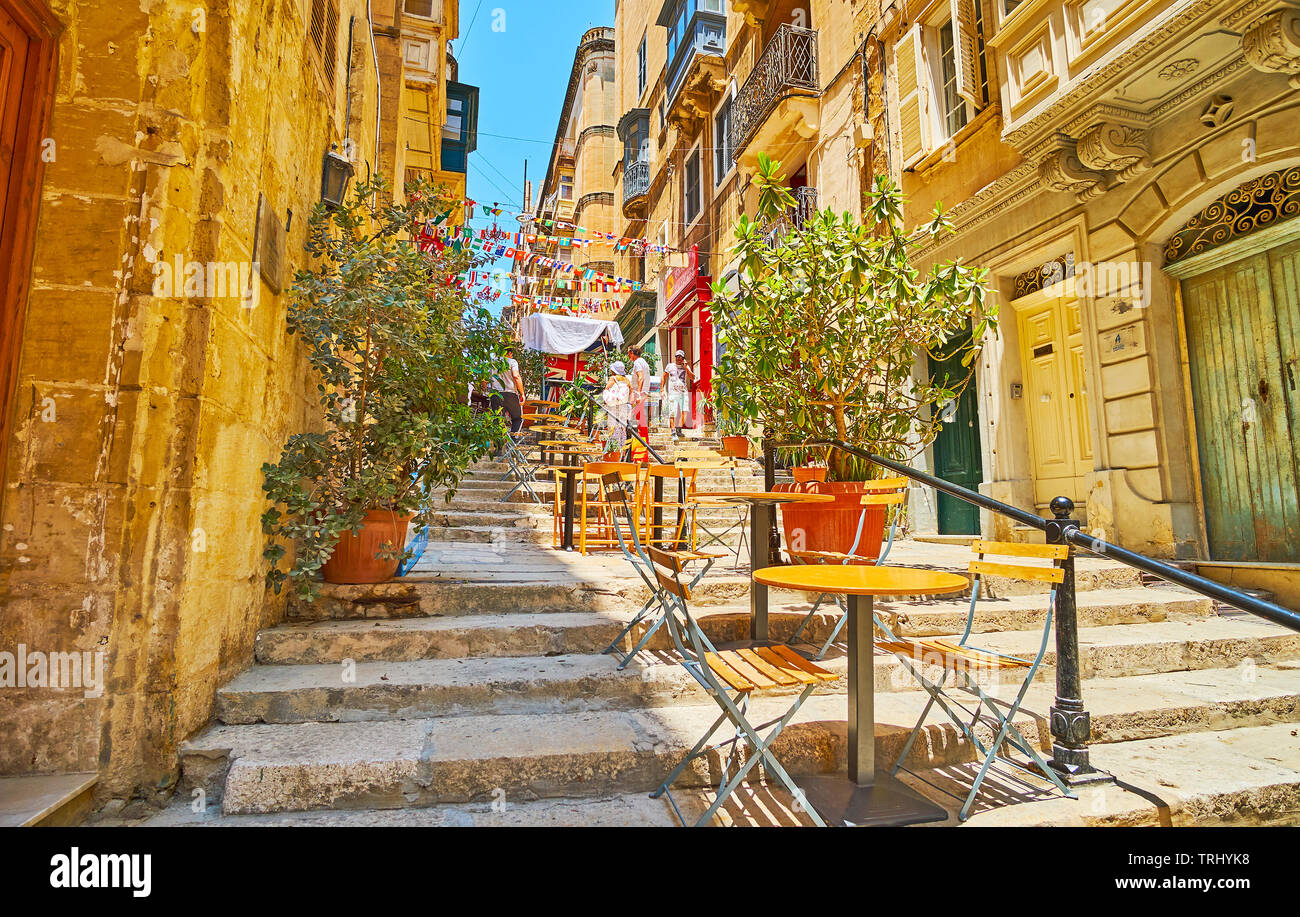 VALLETTA, MALTA - JUNE 19, 2018: The street of St Lucia is old narrow descent, occupied with cozy outdoor cafes and bars, decorated with plants in pot Stock Photo