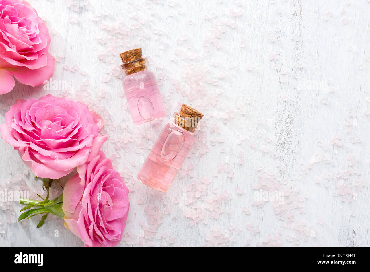 Two bottles with rose oil, crystals of mineral bath salts and pink roses on the wooden table. Stock Photo