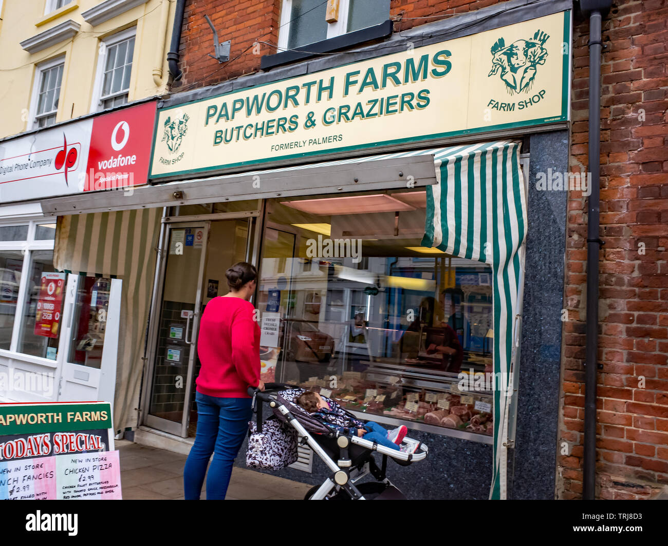 North Walsham, Norfolk, UK. 18 May 2019. Candid street photos – A young mother with her child in a push chair looking at the window display of a high street butcher to choose what meat to buy Stock Photo
