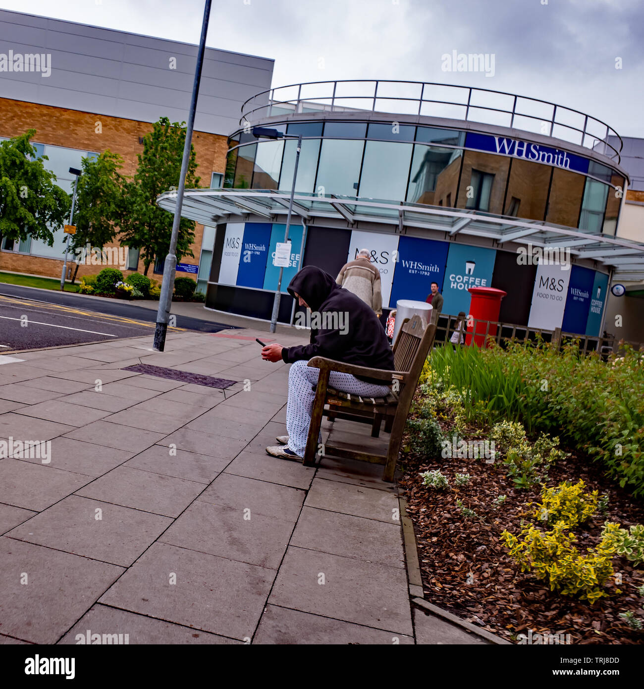 Candid photos - Man in a black hoodie sitting on a wooden bench and using his mobile phone outside at the NHS Norwich University Hospital Stock Photo