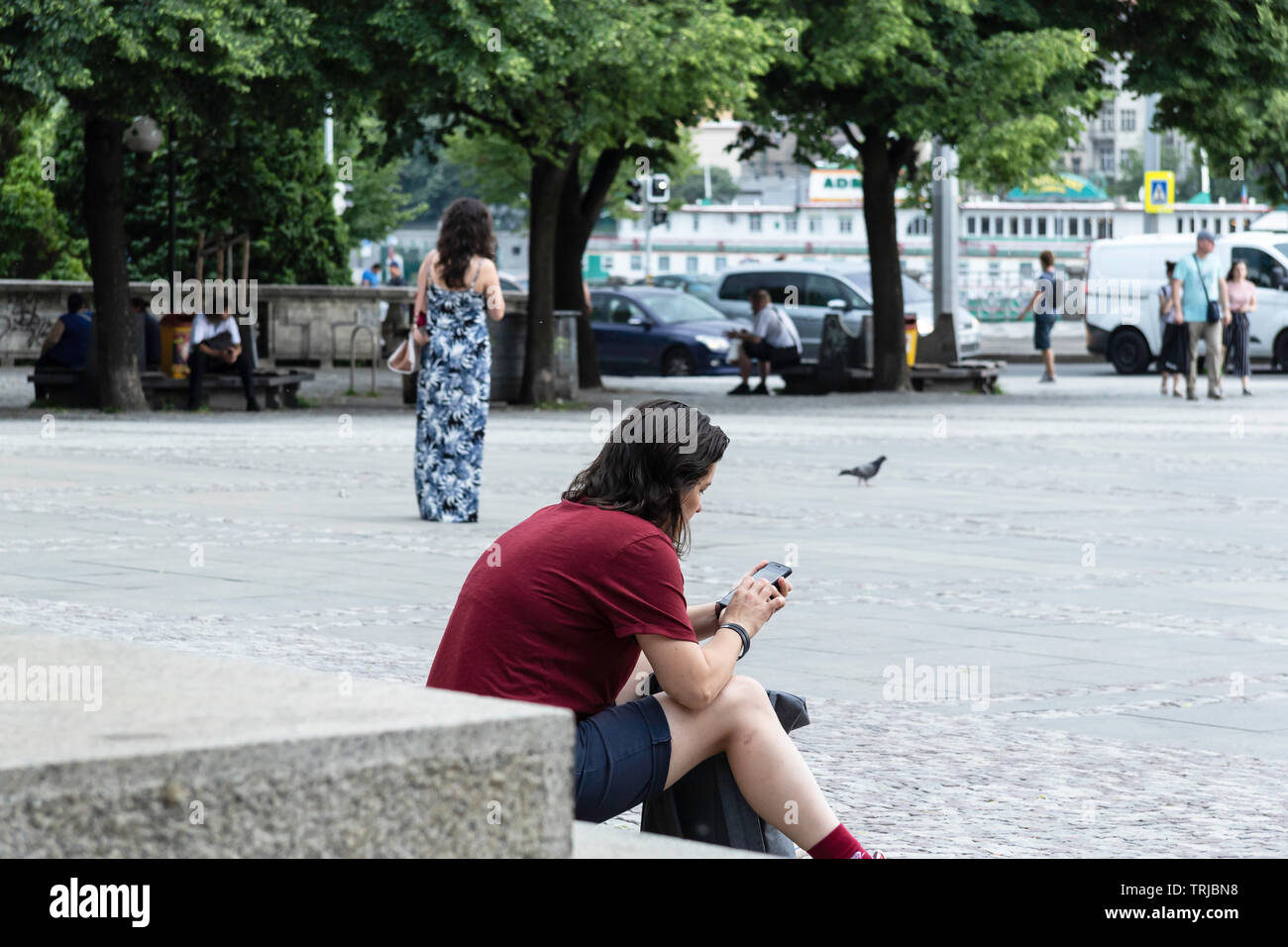 Female sitting on steps at a Square in Prague EU, reading her text messages, background blurred people, trees and cars Stock Photo