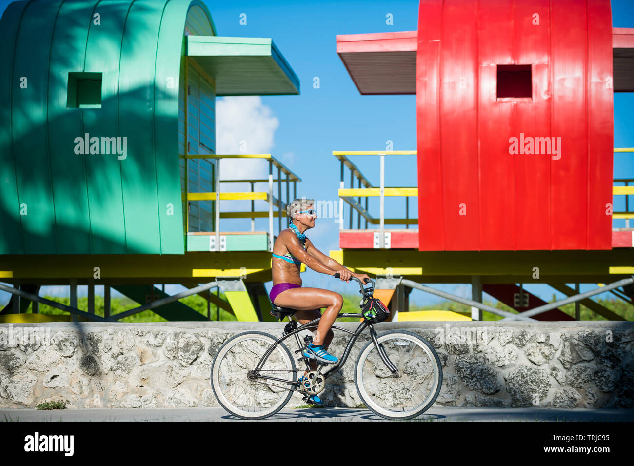 MIAMI - SEPTEMBER, 2018: A mature woman rides a bicycle past a row of brightly colored lifeguard towers awaiting placement on South Beach. Stock Photo