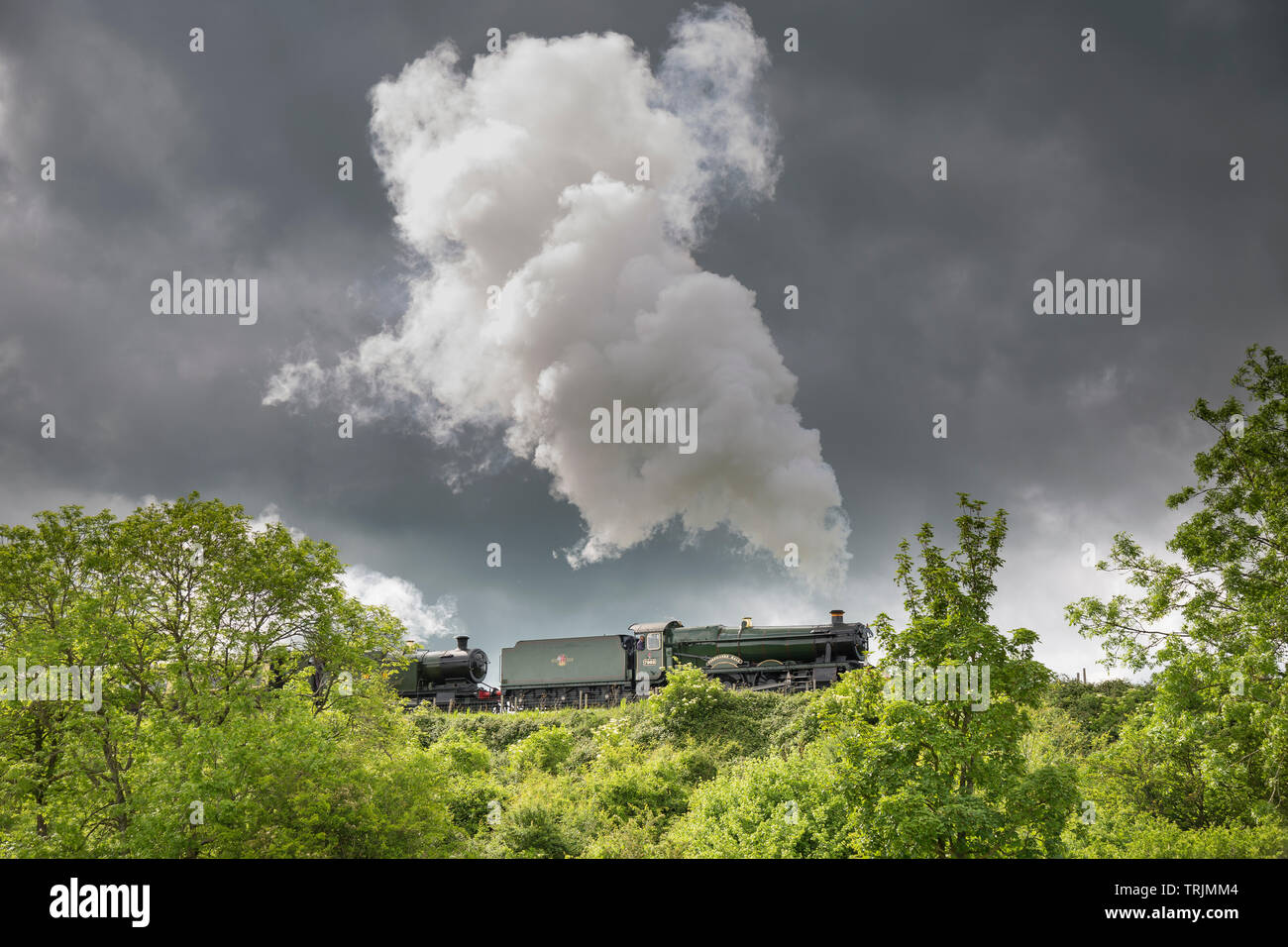 Side view of vintage UK steam train passing through English countryside puffing out steam on GWSR heritage line, sunny afternoon but dark clouds above. Stock Photo
