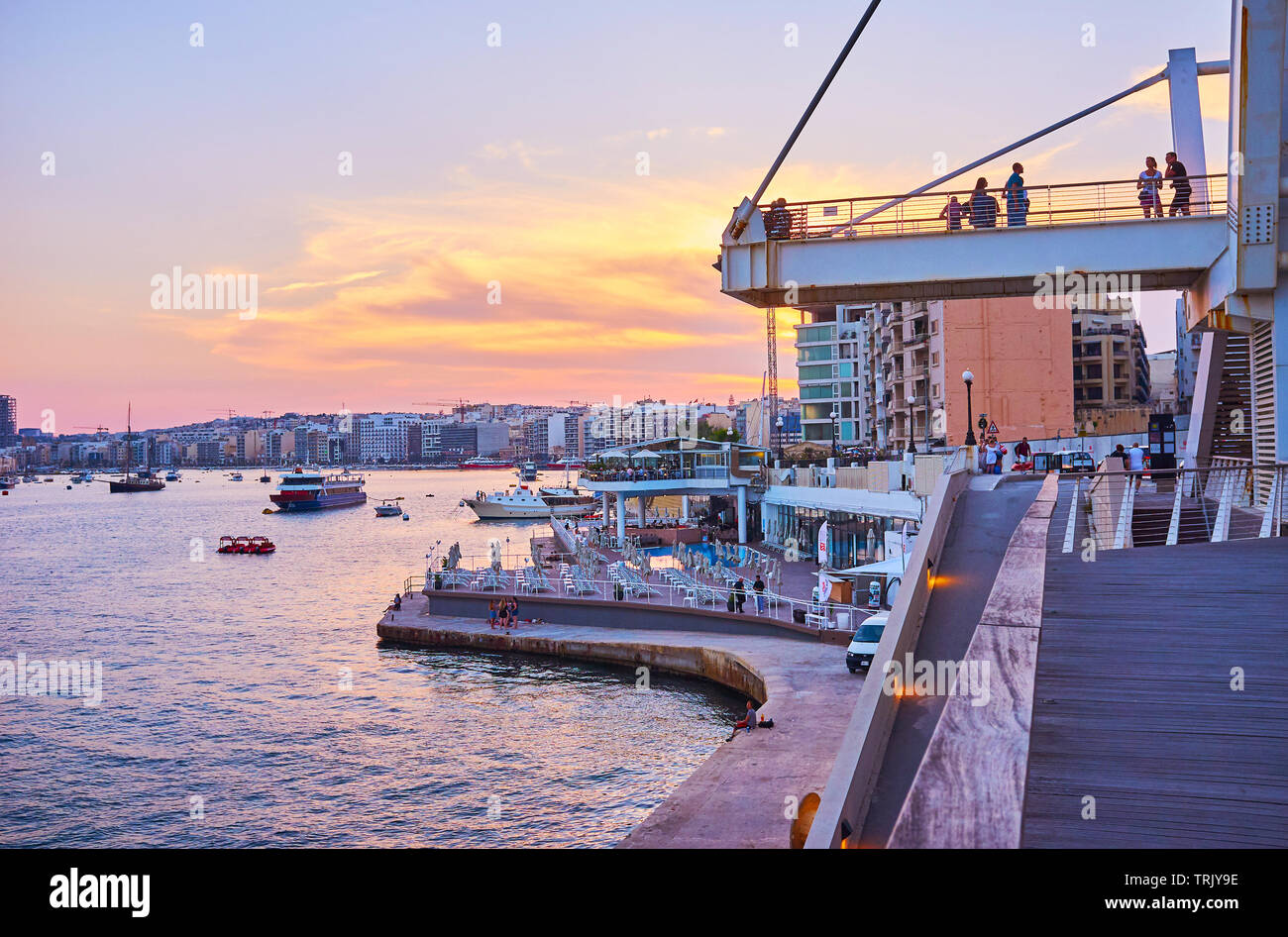 SLIEMA, MALTA - JUNE 19, 2018: The picturesque evening in resort with a view on harbor with ships, fiery sky and comfortable coastal promenade of Tign Stock Photo
