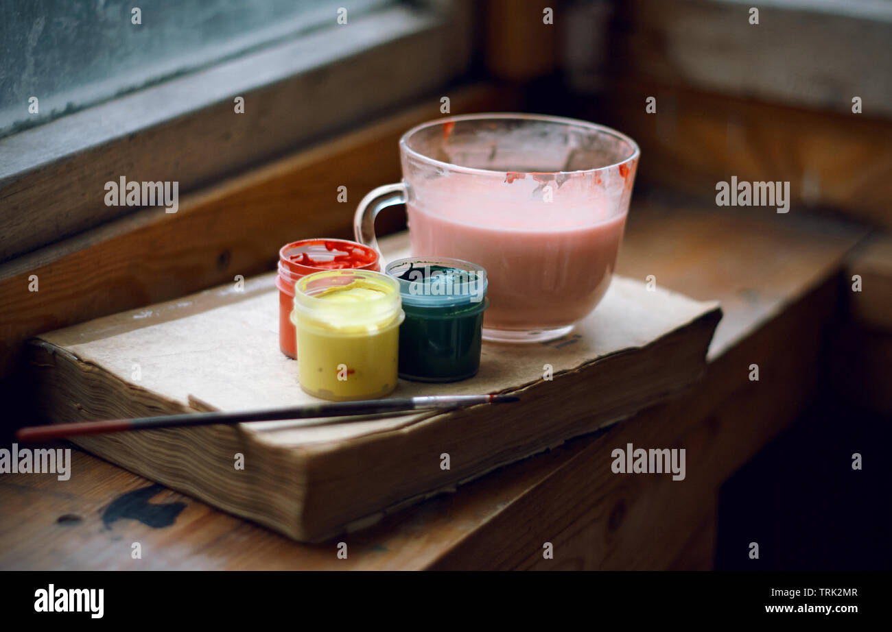 Still life of gouache paints, brushes, paint dilution cup and a book that teaches drawing. All items are on the old wooden windowsill. Stock Photo