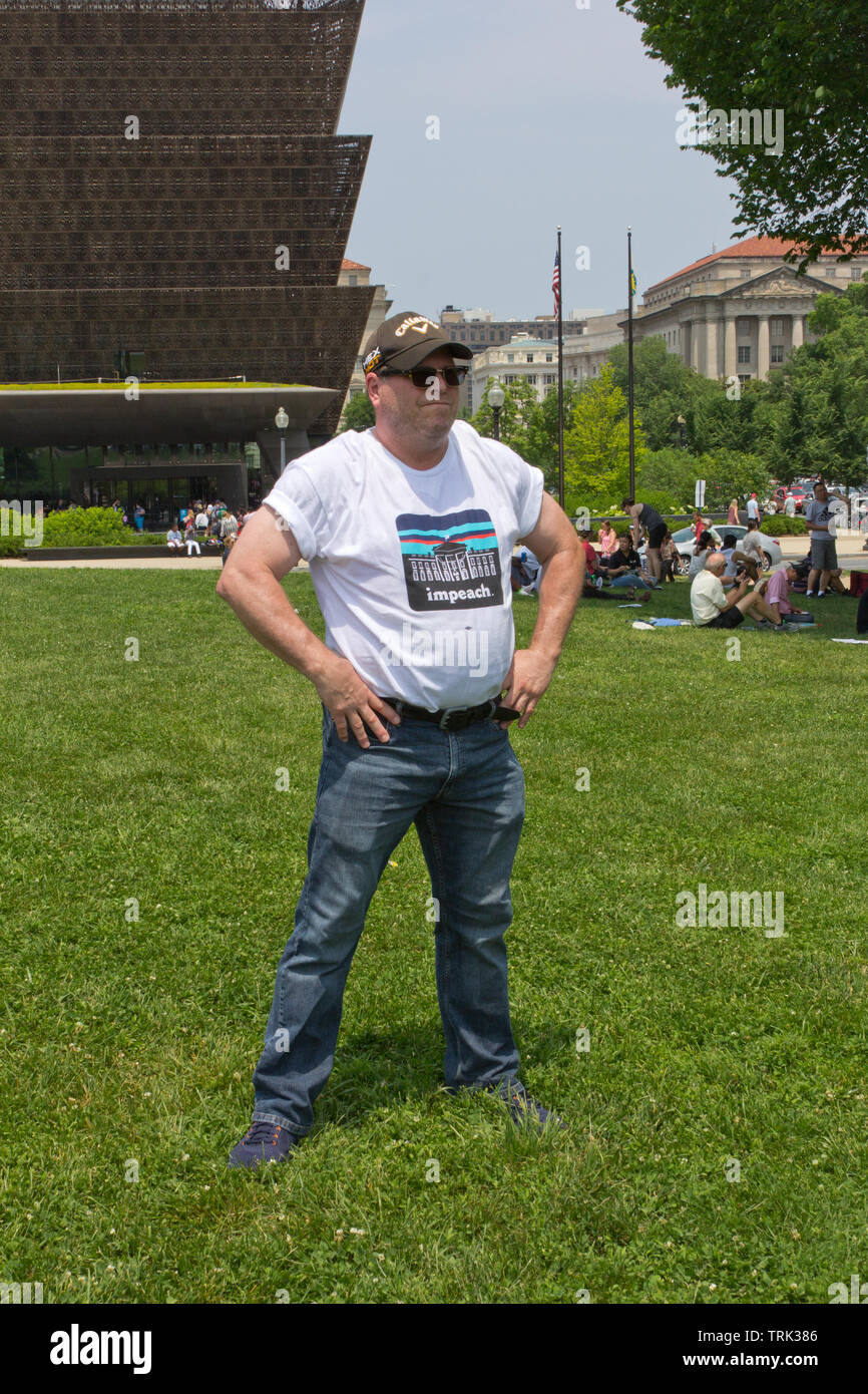 Washington, D.C., USA. 1st June, 2019. With the National Museum of African American History and Culture in the background to the left, a muscular man in akimbo stance wears a t-shirt reading 'Impeach' with a picture of the White House, and a Callaway Hex Hot hat/cap, at the National March To Impeach on 1 June, 2019, on the Washington Monument Grounds in Washington, D.C., USA, organized by People Demand Action. Stock Photo