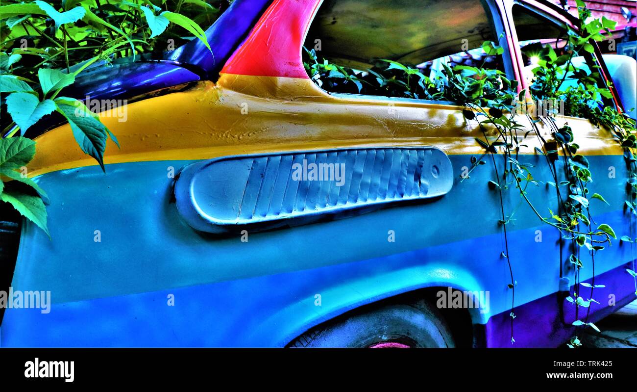 An old Italian Fiat 500 car painted in rainbow colors, submerged in climbing plants, parked on a sidewalk. Stock Photo