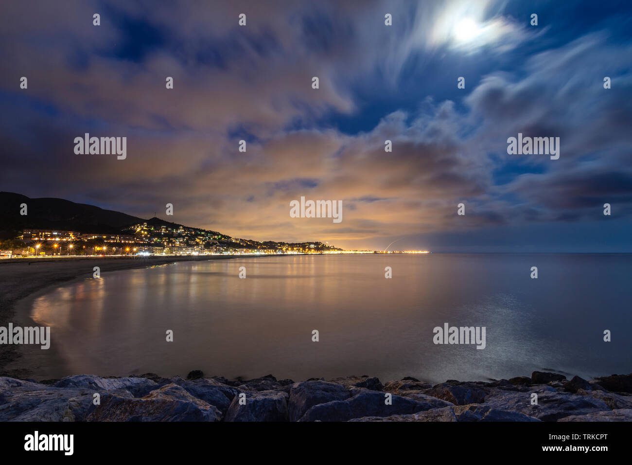 Calm beach at night with spectacular sky and moon, illuminated city background, Catalonia, Spain Stock Photo