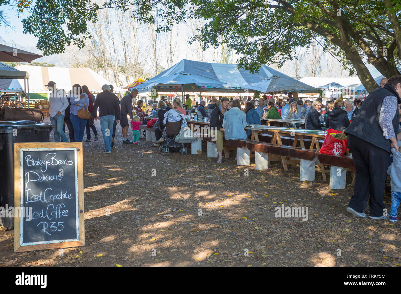 outdoor market venue with people and tourists sitting at benches or tables and chairs under trees at Blaauwklippen wine estate, Stellenbosch Stock Photo