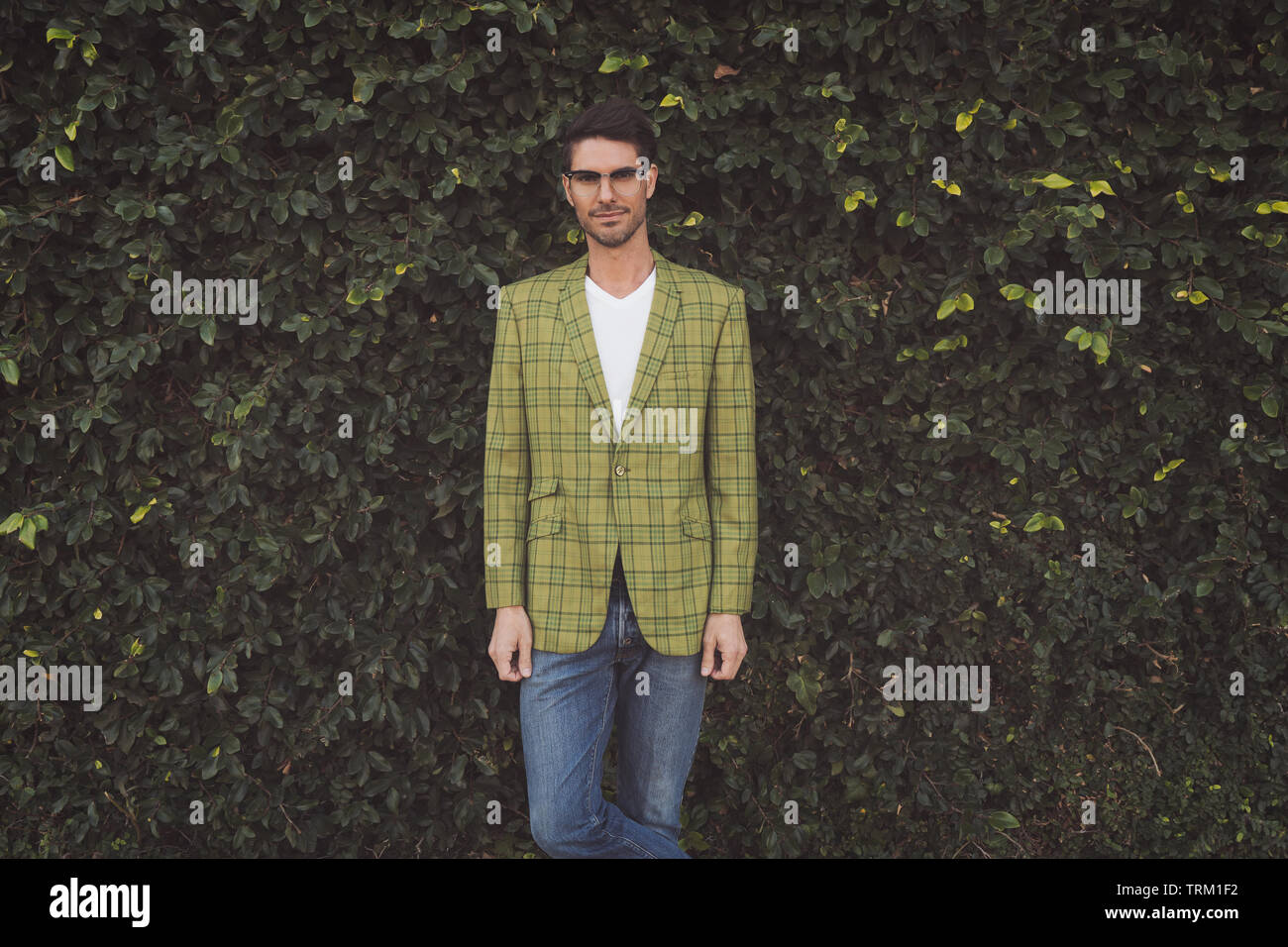 A male model, standing in front of a large green tree bush, looking at the camera, wearing a moss green plaid 70s vintage jacket & vintage eye glasses Stock Photo